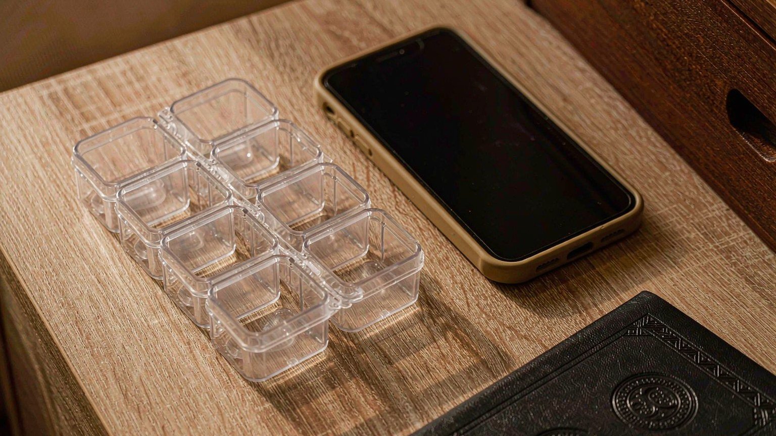A seven-day travel pill organizer beside a phone showing an alarm, set to home time for a trip across time zones